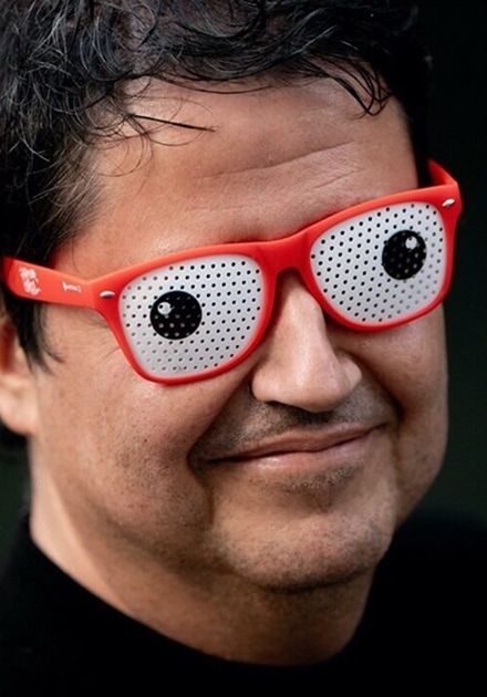 Homem de meia-idade sorri ligeiramente, usando uns óculos de plástico vermelhos com lentes brancas perfuradas que têm grandes olhos desenhados, dando um aspeto cómico ao rosto; o fundo é escuro e desfocado.