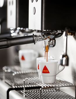 Máquina de café expresso a servir café em chávena branca Delta sobre grelha metálica.