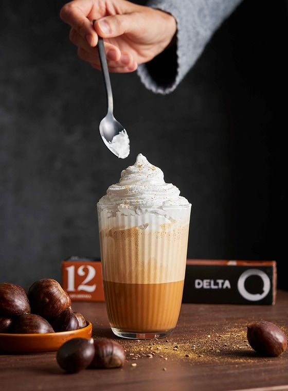 Mão adiciona chantilly a café cremoso em copo de vidro, com castanhas e cápsulas Delta Q na mesa